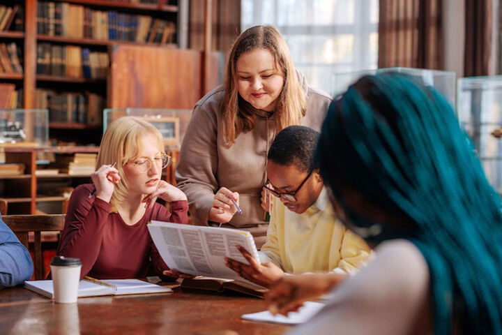 A group of students at a table in a library gather to look at a book together.